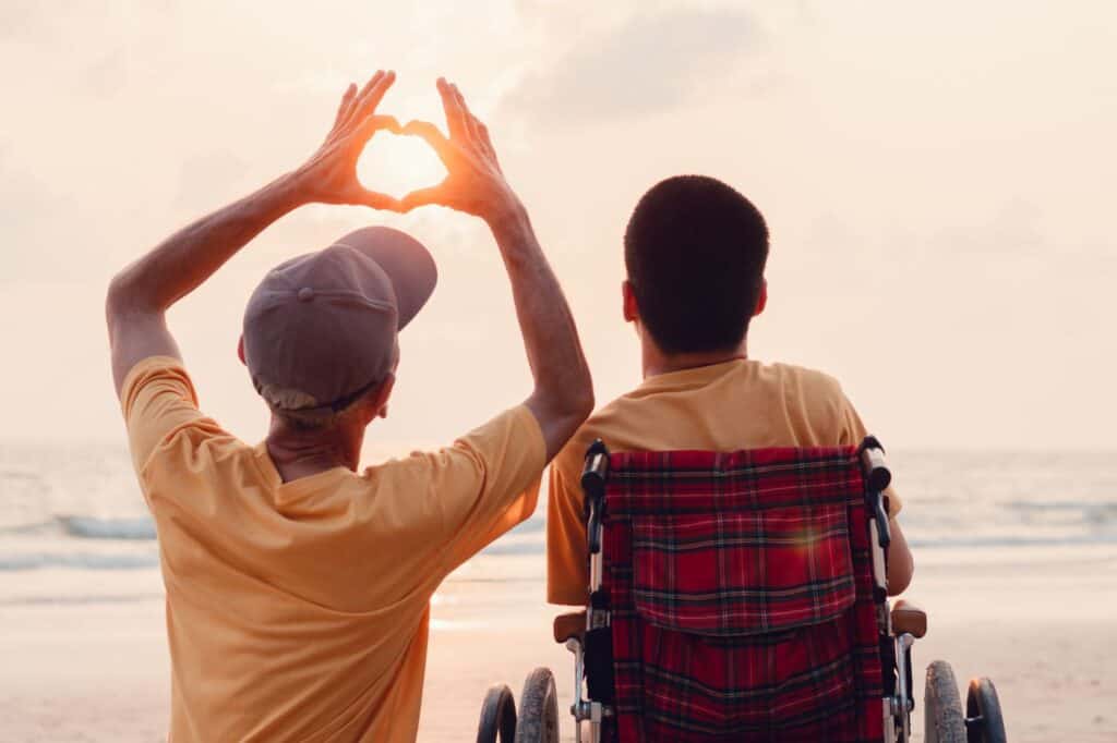 Father and son in a wheelchair making a heart shape at the beach